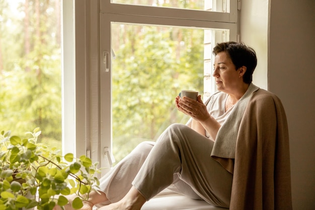 Middle age beautiful woman sitting on windowsill drinking tea dreaming 50yearold woman relaxing with cup of tea Relax at home looking away thinking stress free peaceful mood wellbeing alone