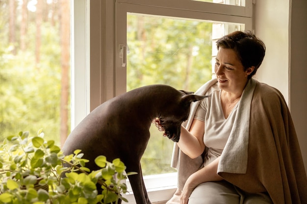그녀의 개와 함께 창턱에 앉아 있는 중년의 아름다운 여성 50세 여성은 집에서 멕시코 털이 없는 개와 시간을 보내고 있습니다.