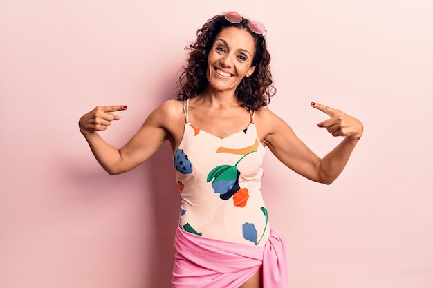 Middle age beautiful woman wearing swimwear and sunglasses looking confident with smile on face pointing oneself with fingers proud and happy