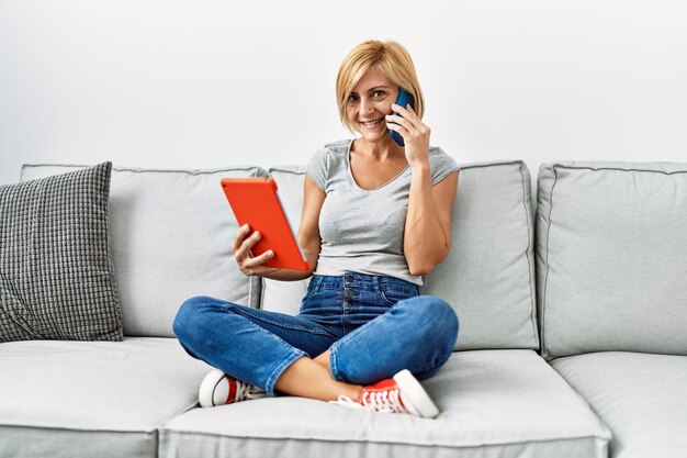 Middle age blonde woman using touchpad and talking on the smartphone at home