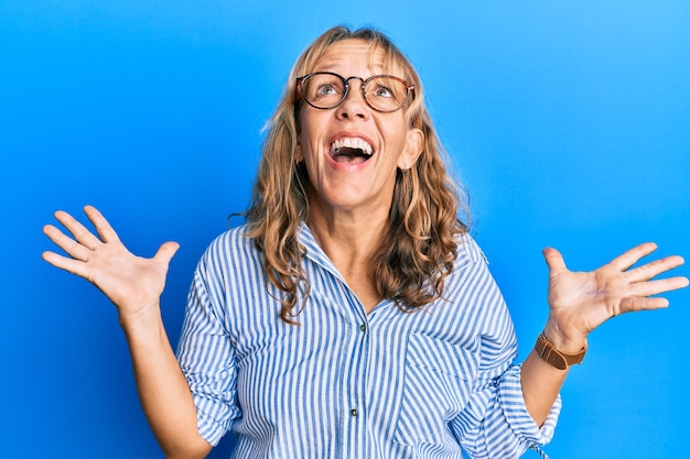 Middle age blonde woman wearing casual clothes and glasses crazy and mad shouting and yelling with aggressive expression and arms raised frustration concept