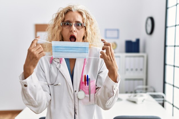 Middle age blonde woman wearing doctor uniform and medical mask afraid and shocked with surprise and amazed expression, fear and excited face.