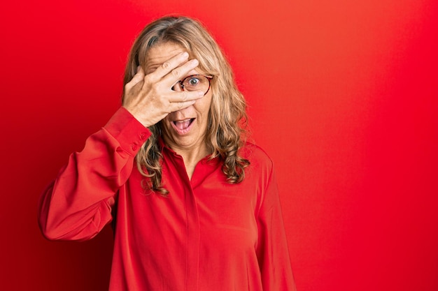 Middle age blonde woman wearing glasses over red background peeking in shock covering face and eyes with hand looking through fingers afraid