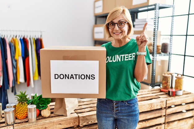 Middle age blonde woman wearing volunteer t shirt holding donations box surprised with an idea or question pointing finger with happy face number one