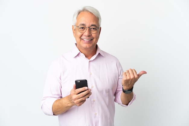 Middle age Brazilian man isolated on white background using mobile phone and pointing to the lateral