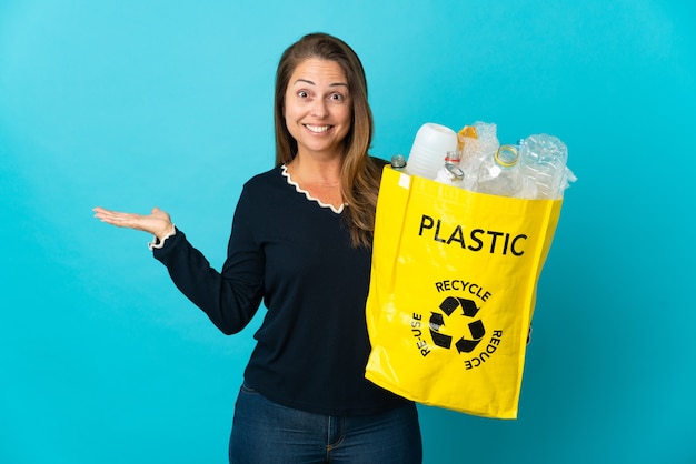 Donna brasiliana di mezza età che tiene un sacchetto pieno di bottiglie di plastica da riciclare sulla parete blu con l'espressione facciale scioccata