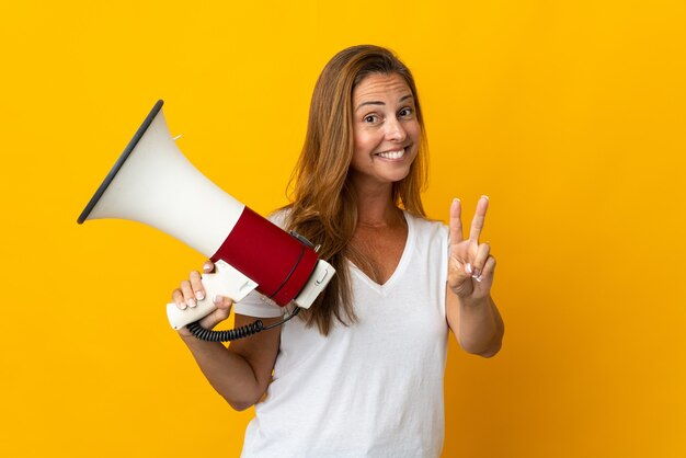 Middle age brazilian woman isolated on yellow holding a megaphone and smiling and showing victory sign