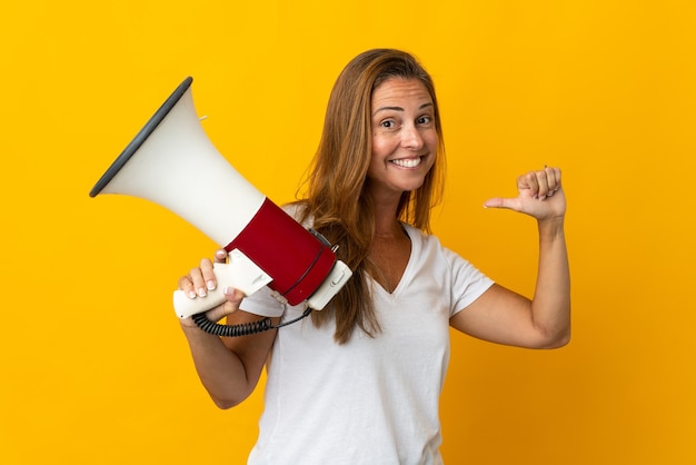 Middle age brazilian woman isolated on yellow wall holding a megaphone and proud and self-satisfied