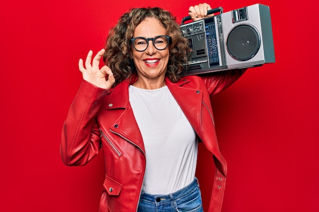 赤い背景にレトロ音楽のブームボックスを握っている中年ブルネットのヒップスターの女性OKをしている指で笑顔で友好的なジェスチャーをしている優れたシンボル