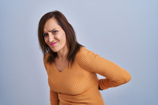 Middle age brunette woman standing wearing orange sweater suffering of backache, touching back with hand, muscular pain