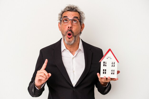 Middle age business man holding bills and house model isolated on blue background  pointing upside with opened mouth.