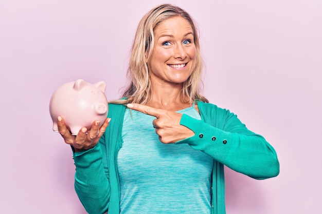 Middle age caucasian blonde woman holding piggy bank smiling happy pointing with hand and finger