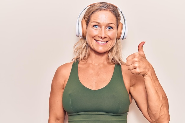 Middle age caucasian blonde woman listening to music using headphones smiling happy and positive thumb up doing excellent and approval sign