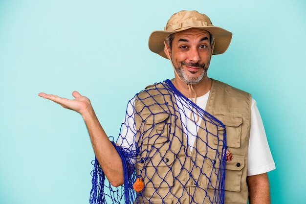 Middle age caucasian fisherman holding net isolated on blue background  showing a copy space on a palm and holding another hand on waist.