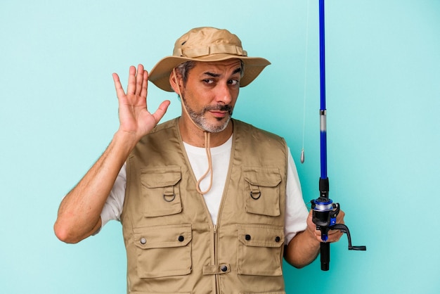 Middle age caucasian fisherman holding rod isolated on blue background trying to listening a gossip