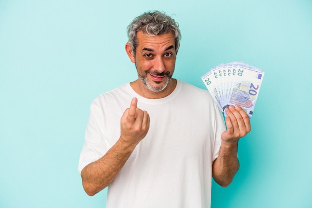 Middle age caucasian man holding bills isolated on blue background  pointing with finger at you as if inviting come closer.