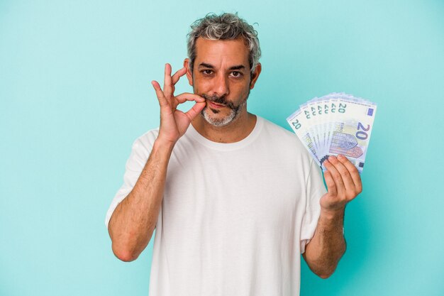 Middle age caucasian man holding bills isolated on blue background  with fingers on lips keeping a secret.