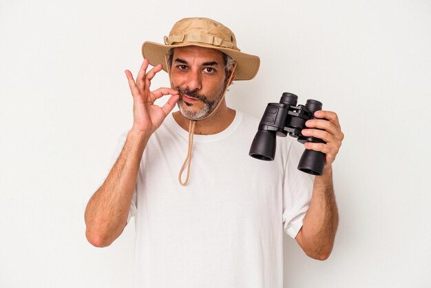 Middle age caucasian man holding binoculars isolated on white\
background with fingers on lips keeping a secret