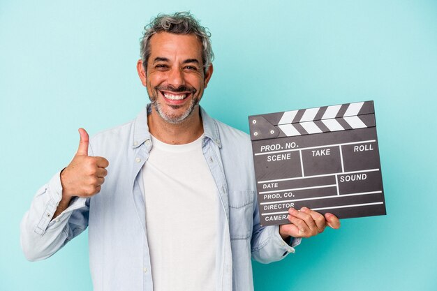 Middle age caucasian man holding a clapperboard isolated on blue background  smiling and raising thumb up