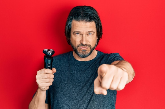 Photo middle age caucasian man holding electric razor machine pointing with finger to the camera and to you, confident gesture looking serious