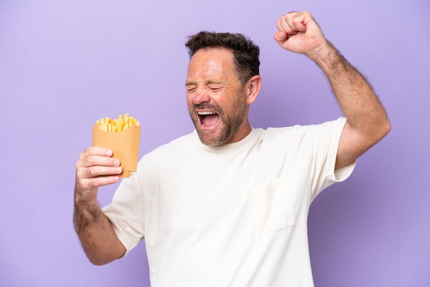 Photo middle age caucasian man holding fried chips isolated on purple bakcground celebrating a victory