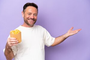 Middle age caucasian man holding fried chips isolated on purple bakcground extending hands to the side for inviting to come