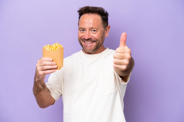 Middle age caucasian man holding fried chips isolated on purple bakcground with thumbs up because something good has happened