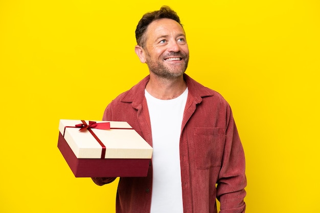 Middle age caucasian man holding a gift isolated on yellow background looking up while smiling