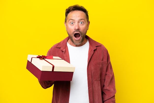 Middle age caucasian man holding a gift isolated on yellow background with surprise and shocked facial expression