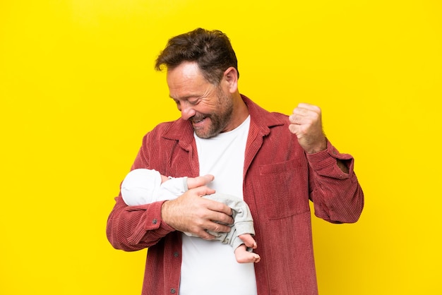 Middle age caucasian man holding his newborn son isolated on yellow background celebrating a victory