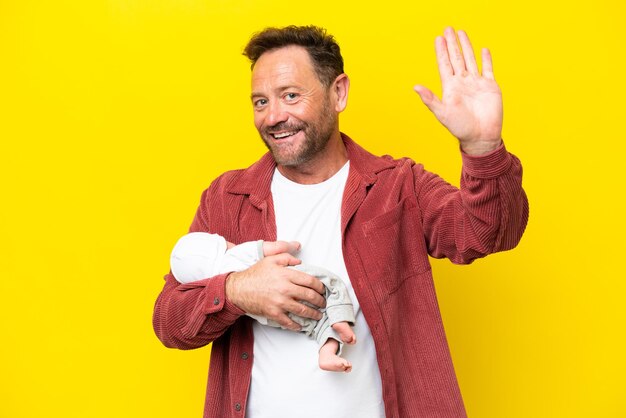 Middle age caucasian man holding his newborn son isolated on yellow background saluting with hand with happy expression