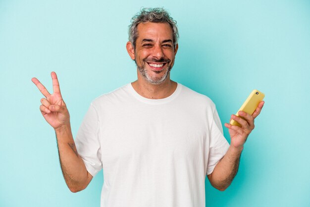 Middle age caucasian man holding a mobile phone isolated on blue background  joyful and carefree showing a peace symbol with fingers.