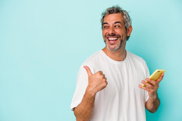 Middle age caucasian man holding a mobile phone isolated on blue background  points with thumb finger away, laughing and carefree.