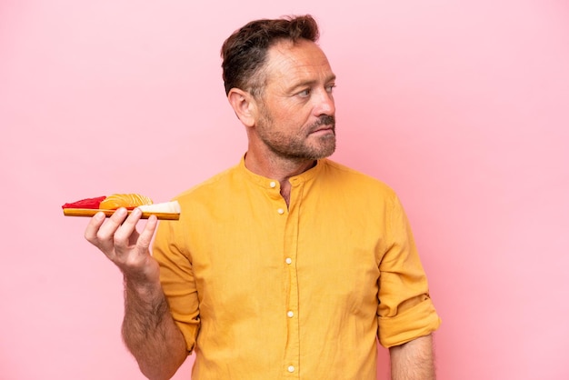 Middle age Caucasian man holding sashimi isolated on pink background looking to the side