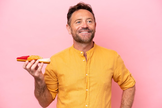 Middle age Caucasian man holding sashimi isolated on pink background posing with arms at hip and smiling