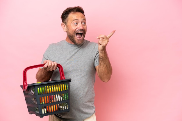 Middle age caucasian man holding a shopping basket isolated on pink background intending to realizes the solution while lifting a finger up