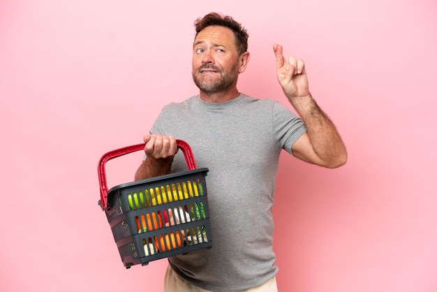 Middle age caucasian man holding a shopping basket isolated on pink background with fingers crossing and wishing the best