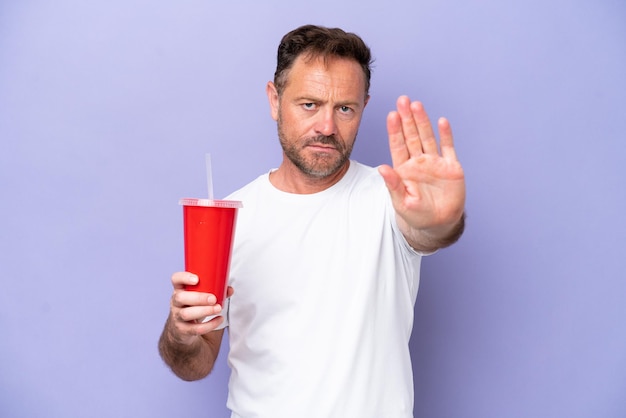 Middle age caucasian man holding soda isolated on purple background making stop gesture