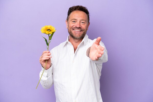 Middle age caucasian man holding sun flower isolated on purple background shaking hands for closing a good deal