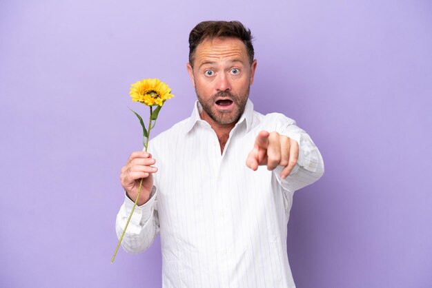 Middle age caucasian man holding sun flower isolated on purple background surprised and pointing front