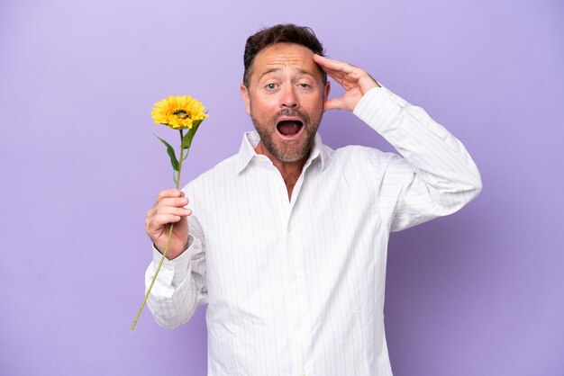 Middle age caucasian man holding sun flower isolated on purple background with surprise expression