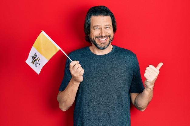 Middle age caucasian man holding vatican city flag pointing thumb up to the side smiling happy with open mouth