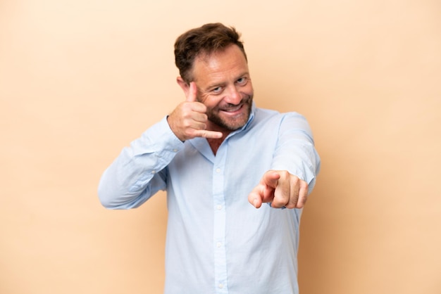 Foto uomo caucasico di mezza età isolato su sfondo beige che fa il gesto del telefono e punta davanti