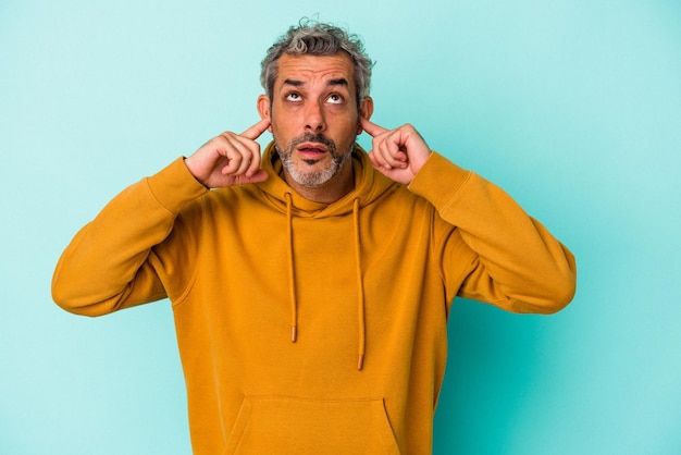 Middle age caucasian man isolated on blue background  covering ears with fingers, stressed and desperate by a loudly ambient.