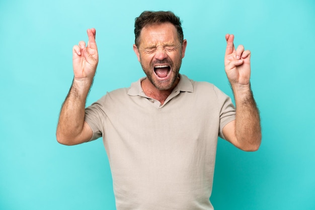 Middle age caucasian man isolated on blue background with fingers crossing