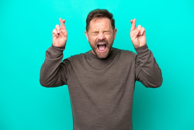 Middle age caucasian man isolated on blue background with fingers crossing