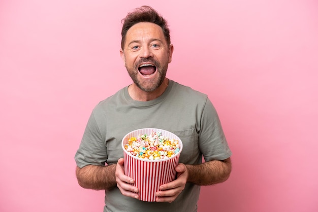 Middle age caucasian man isolated on pink background holding a big bucket of popcorns