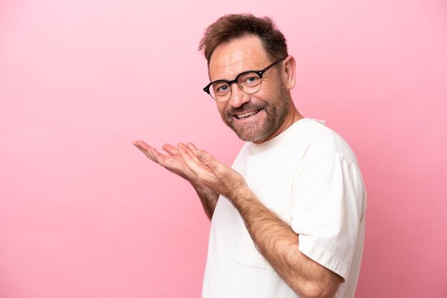 Middle age caucasian man isolated on pink background With glasses and presenting something