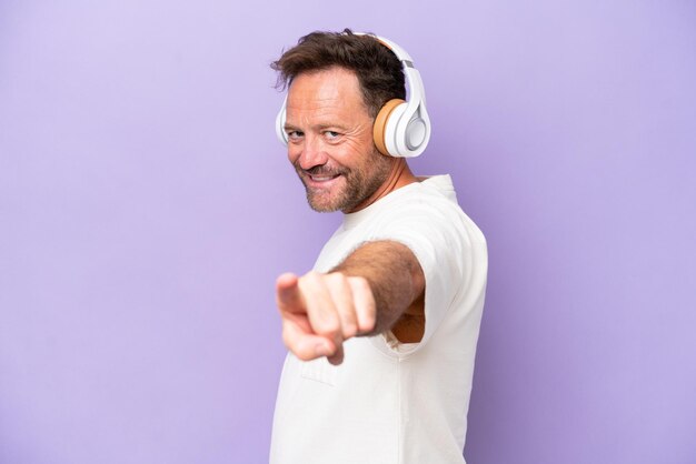 Middle age caucasian man isolated on purple background listening music and pointing to the front