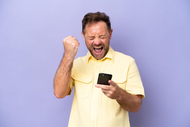 Middle age caucasian man isolated on purple background with\
phone in victory position